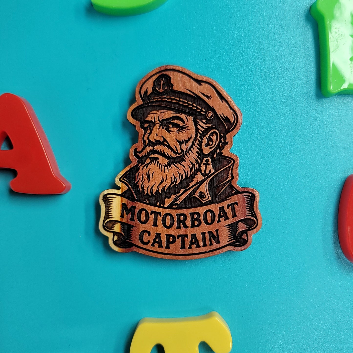 A brown and gold metal pin shaped like a captain's portrait with the words 'MOTORBOAT CAPTAIN' on a banner, displayed on a bright blue background with colorful letter magnets around it.