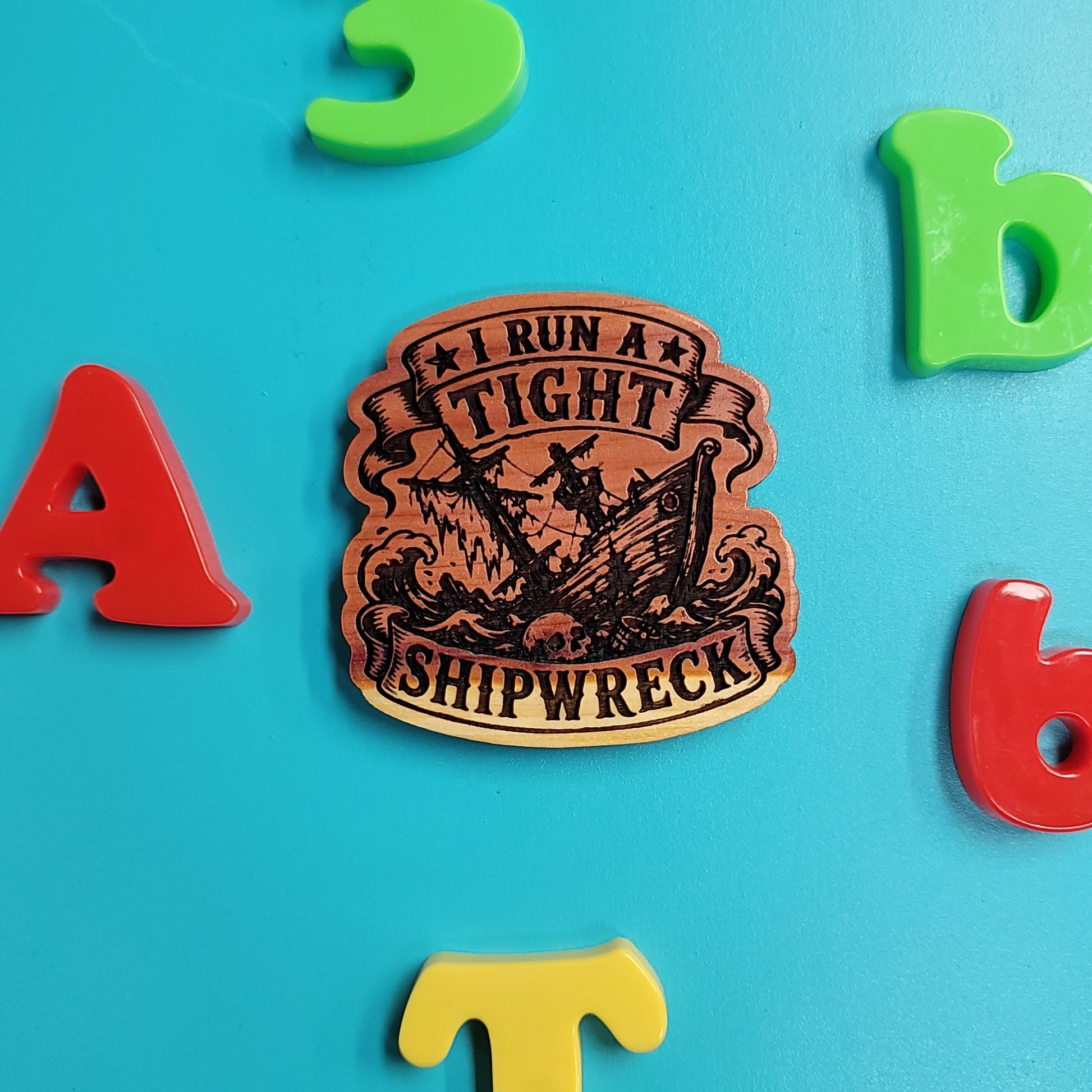 A wooden shipwreck letterpress printing block with black engraved text and illustration, surrounded by colorful plastic alphabet letters on a bright blue background.