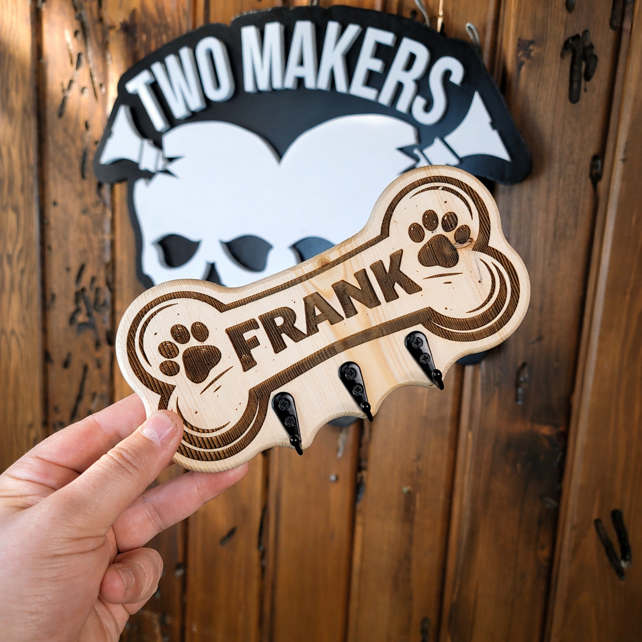 Wooden bone-shaped pet tag with 'FRANK' engraved, held in front of a wooden background with 'TWO MAKERS' logo.